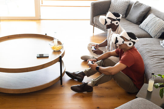 Diverse male friends sitting on hardwood floor in living room wearing VR gear holding controllers