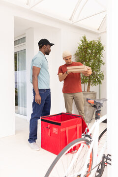 Diverse male delivery worker holding brown boxes while customer watching at porch with red bag