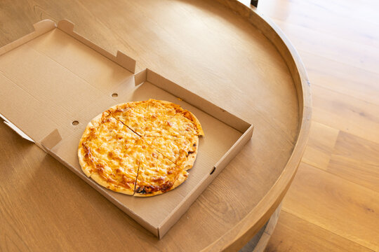 Thin crust cheese pizza sitting in open cardboard box on round wooden table, floor visible