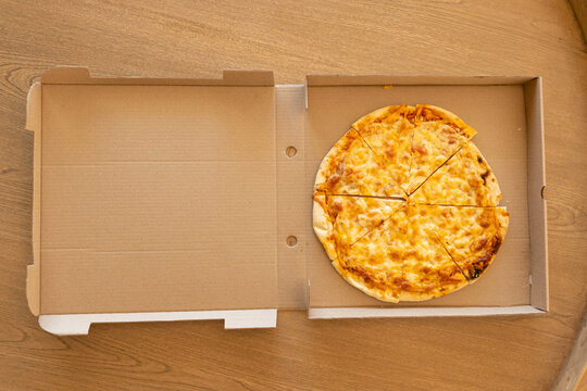 Round cheese pizza is resting in open corrugated box on wooden tabletop, showing charred crust