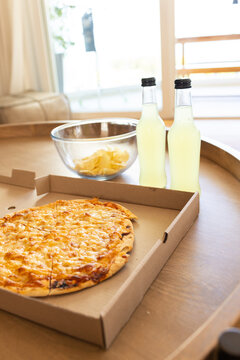 Whole cheese pizza sitting on round wooden table near bright window, with bottles and chips