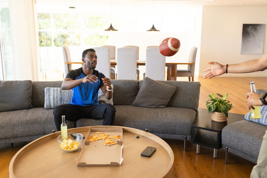 Mid adult male friends wearing athletic shirts catching football on gray sectional sofa with pizza