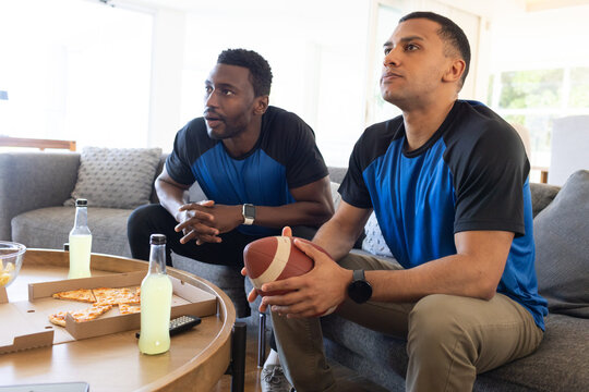 Diverse male friends in matching shirts watching game at home holding football pizza, copy space
