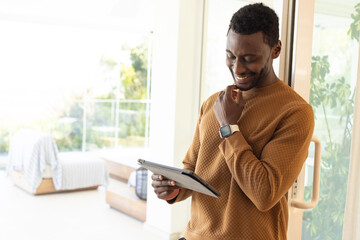 Mid adult African American man leaning on glass door holding tablet, smartwatch at home, copy space