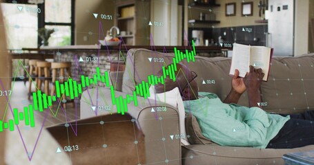 Reclining senior man in green shirt, holding paperback, reading on sofa at home with finance charts