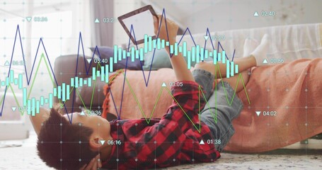 Lying Chinese boy holding tablet on rug at home, wearing checkered shirt, denim shorts, with charts