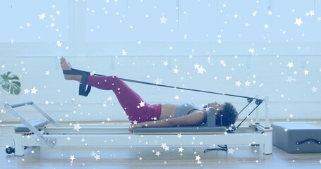 Lying woman wearing gray sports bra, magenta leggings, pulling straps on reformer in studio, plant