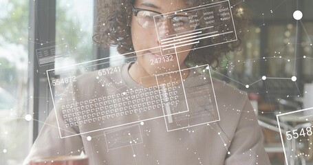 Working curly-haired man with glasses using holographic interface on tablet by cafe window, sweater