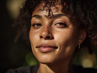 Smiling person with curly hair in close-up, looking confidently at the camera. Soft lighting highlights facial features and subtle freckles. Golden hoop earrings are visible
