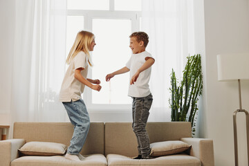 Two happy children jumping on beige sofa in bright living room with large window and green plant....