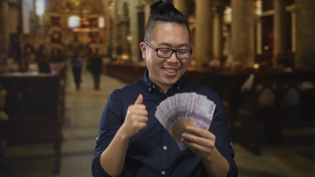 Young man holding indonesian rupiah banknotes and giving thumbs up in catholic church building; joy prosperity.