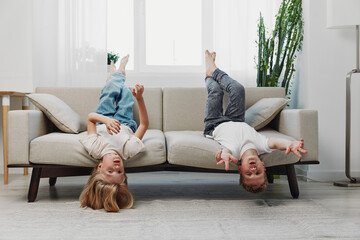 Two happy children hang upside down from a beige sofa in a bright living room with plants, enjoying playful moments and laughter. Concept of childhood fun and carefree home environment. © SHOTPRIME STUDIO