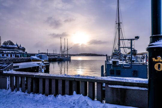 Harbor view in Oslo, Norway, with boats, people in silhouette, winter sunlight reflecting on the sea during a snowy sunset.