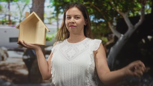 Woman holding wooden house model with left hand and points finger at camera in building; homeownership confidence.