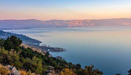 A scenic coastal view showcasing a tranquil body of water, framed by a lush foreground of trees and distant mountain ranges under a pastel sky