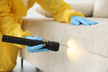 Pest control worker in protective suit examining sofa with flashlight indoors, closeup