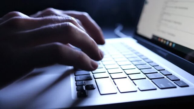 Close-up of male hands typing on laptop keyboard at night working late in dark room