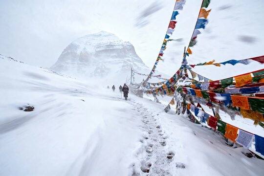 Snow-covered mountain path lined with colorful prayer flags, with hikers ascending toward a distant peak in a misty, high-altitude landscape. - Powered by Adobe