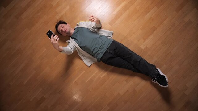 Man recording video or taking a selfie, lying casually on a light wooden floor from a high angle view, creating content for social media with his mobile device