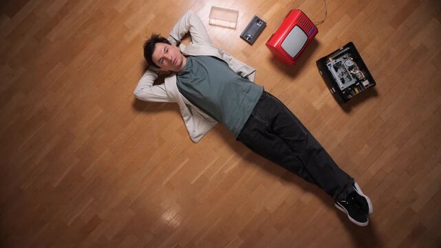 Man lying on a wooden floor, surrounded by vintage electronic devices like a small television and an old cassette player, creating a sense of leisure and nostalgia for classic media