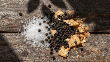 Coarse salt and black peppercorns are scattered with broken crackers on a wooden surface