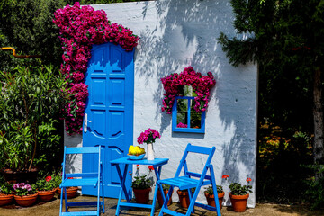 The photograph shows a dummy house with white walls, chairs and an entrance door with a blue window and beautiful red flowers hanging down.