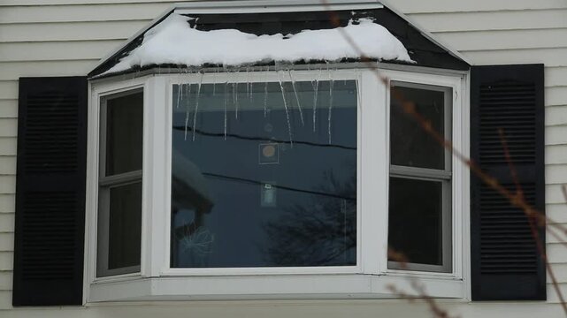 Snow falls in front of a bay window with icicles