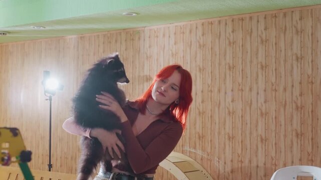 Caucasian woman cuddling fluffy dog against woodpaneled wall, red hair lit by studio lamp, joyful smile while lifting black puppy, warm domestic atmosphere, candid pet owner moment filled