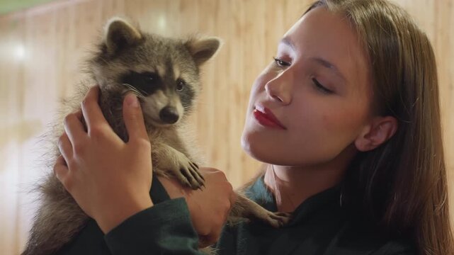 caucasian woman holding raccoon indoors, tender rehabilitator bonding with gentle kisses and soft paw touches, warm wooden interior, calm evening light, affectionate rescue care and playful trust