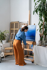 Skillful focused female artist painting picture on canvas at home art studio, adjusting easel...