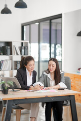 Obraz premium Two professional Asian businesswomen are engaged in a productive discussion at a wooden office desk. One woman is pointing at a laptop screen while the other listens attentively, surrounded by financi