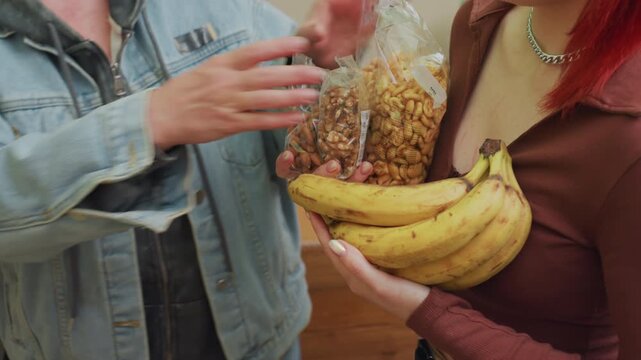 woman holds bananas and granola bag in cozy bulk shop as shopper in denim jacket transfers cereal, closeup hands show barter and teamwork between customer and vendor, warm tones, reflective