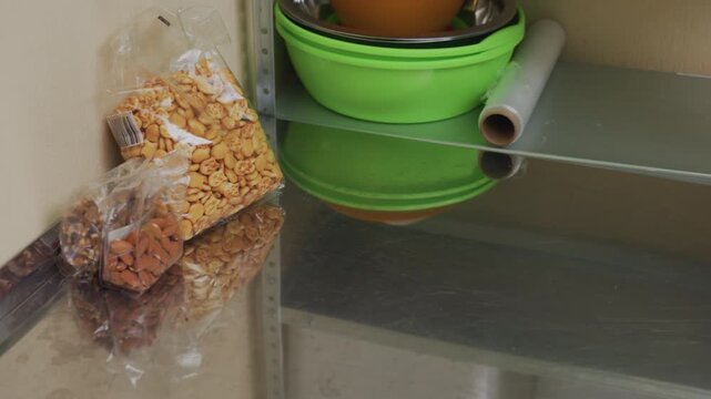 hands place bananas then remove granola in compact storage area where denim jacket hands arrange produce and clear packaging, reflective metal countertop, green bowls on shelf, methodical restock