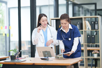 Obraz premium Two female healthcare workers, one in blue scrubs and another in a white lab coat, are collaborating and reviewing patient charts on a clipboard and tablet. They are engaged in a professional discussi