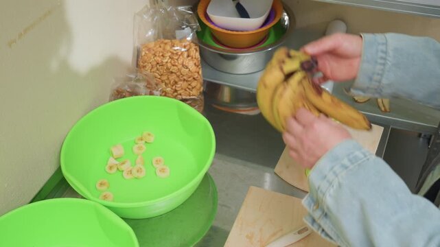 green bowls on stainless counter, bananas and cereal hands reach for bunch beside cutting board and knife, tidy pantry items and scale visible, cozy morning meal prep with organic fruit and simple