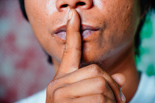 A couple sits on the bed with their backs to each other. A man demonstrates a finger gesture in front of his lips which symbolizes an instruction to be quiet or keep a secret.