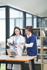 Fototapeta premium Two female healthcare workers, one in blue scrubs and another in a white lab coat, are collaborating and reviewing patient charts on a clipboard and tablet. They are engaged in a professional discussi