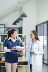 Obraz premium female doctor in a white lab coat and a medical assistant in blue scrubs are sitting at a wooden desk, collaborating and reviewing medical documents or X-rays. Both are wearing stethoscopes, reflectin