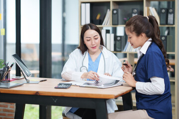 Obraz premium female doctor in a white lab coat and a medical assistant in blue scrubs are sitting at a wooden desk, collaborating and reviewing medical documents or X-rays. Both are wearing stethoscopes, reflectin