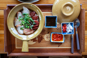 Traditional clay pot baked rice topped with sliced BBQ pork, Chinese sausage, and fresh coriander. Served with spicy dipping sauces on a wooden tray, featuring authentic Asian fusion cuisine.