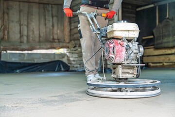 A focused construction worker in red gloves operates a power trowel to smooth a freshly poured...