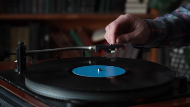 Close-up of a hand gently placing the tonearm onto a spinning vinyl record in a warm, book-filled room. The scene evokes a nostalgic connection to analog music, enhanced by the casual plaid shirt and