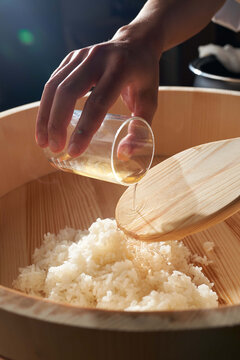 Chef pouring seasoned rice vinegar into a traditional wooden hangiri bowl with steamed white rice for sushi preparation.