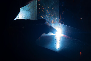 Manual metal welding, welding work in a mask, welding arc, sparks in the dark, bright flash of a welding arc, industrial welding in a protective suit, welding mask