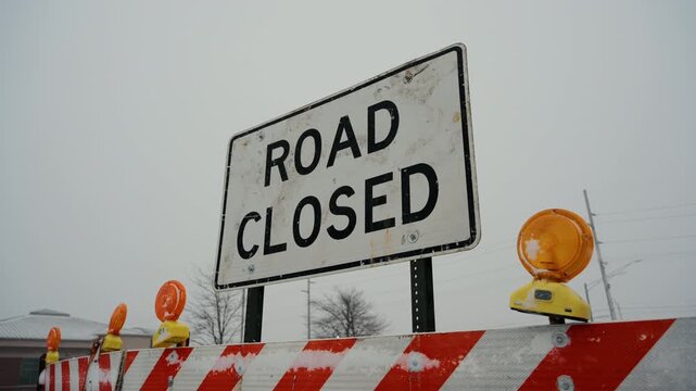 Road closed sign stand behind road closure barriers during snowy winter conditions. traffic restrictions, blocked access due to weather or road work. 