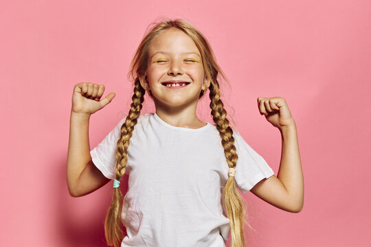 Happy girl with braided hair smiles showing missing teeth while flexing arms on pink background.