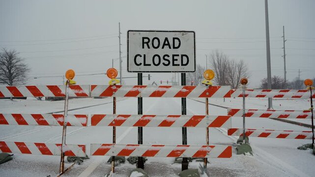 Road closed sign stands behind road closure barriers during snowy winter conditions. traffic restrictions, blocked access due to weather or road work. 
