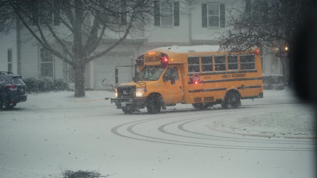 Yellow school bus stopping at suburban neighborhood during active snowfall. Snow blankets the street as flakes fall steadily, creating cold winter scene POV View