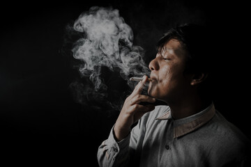 A man smoking a cigarette in the dark represents the dangers and risks of tobacco and nicotine. A man hiding and finding relief from bad habits, stress, and addiction against a black studio background