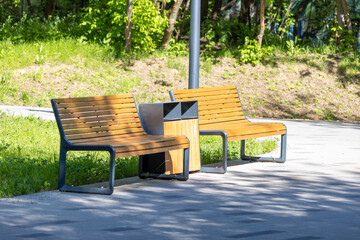 Wooden benches beside a trash bin in a sunny park, surrounded by lush green trees and grass, casting shadows on the paved pathway in a serene outdoor setting